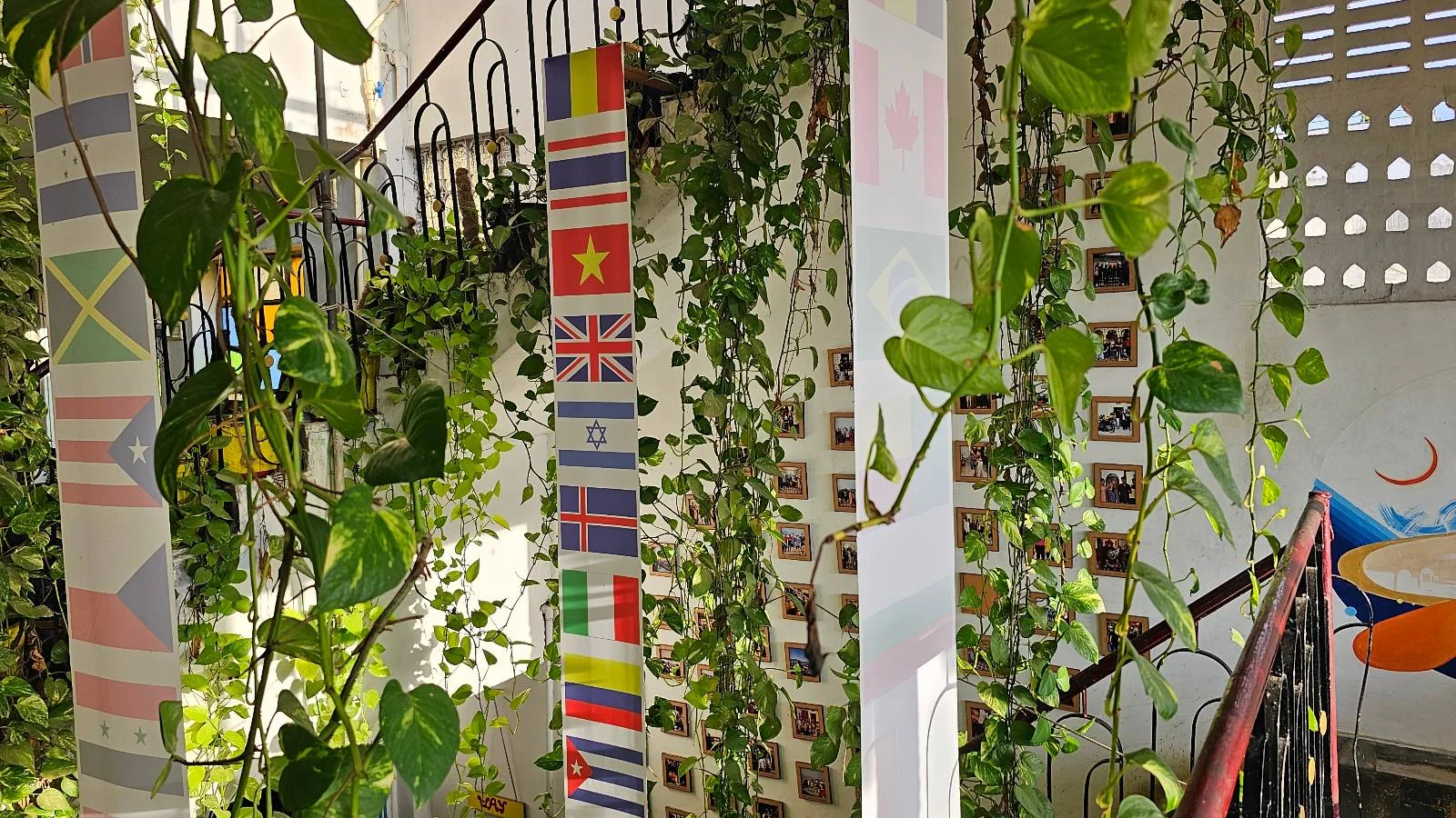 Indoor staircase surrounded by lush green plants, with several white columns decorated with colorful world flags. Sunlight filters through the space, creating a bright and vibrant atmosphere.