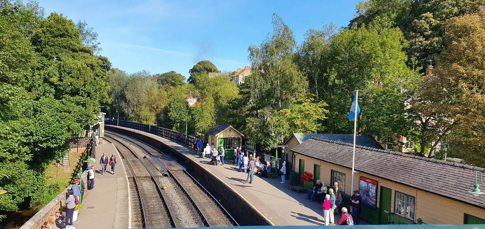A sunny day at a small train station with people waiting on the platform, surrounded by trees and greenery; a yellow building stands to the right, with train tracks curving into the distance.