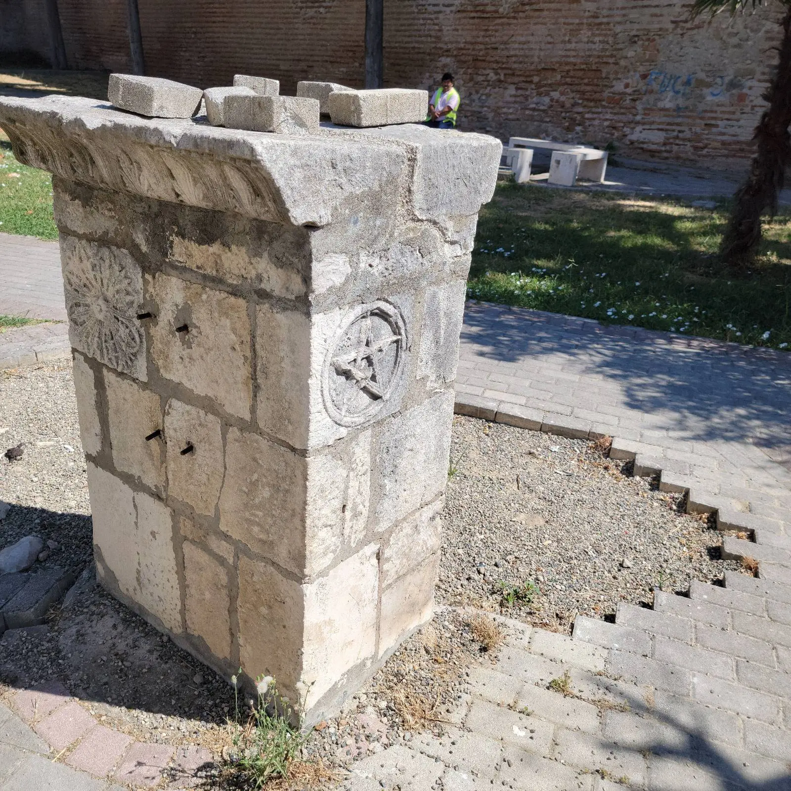 Stone pedestal with engraved symbols in a sunny park. Background shows a person sitting on a bench against a brick wall. Grass and shadows.