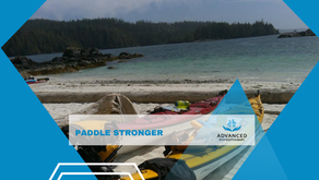 Kayaks on a sandy beach with a forested shoreline. Text reads "PADDLE STRONGER" and "ADVANCED PHYSIOTHERAPY." Calm, outdoor scene.