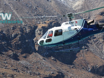 Blue and white helicopter flying over rocky mountainous terrain. Marked 9N-AKB and "Mountain." Clear blue sky. Logo: rw networks.