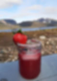 A glass jar filled with a deep red beverage sits on a wooden surface, garnished with a sugared rim and a fresh, ripe strawberry perched on top. In the background, a serene landscape unfolds with a calm lake, rocky terrain, and majestic mountains partially dusted with snow beneath a partly cloudy sky.