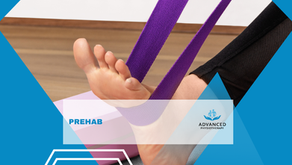 Foot stretches with a purple band on a yoga mat. "PREHAB" text and "ADVANCED PHYSIOTHERAPY" logo are visible. Wood floor background.