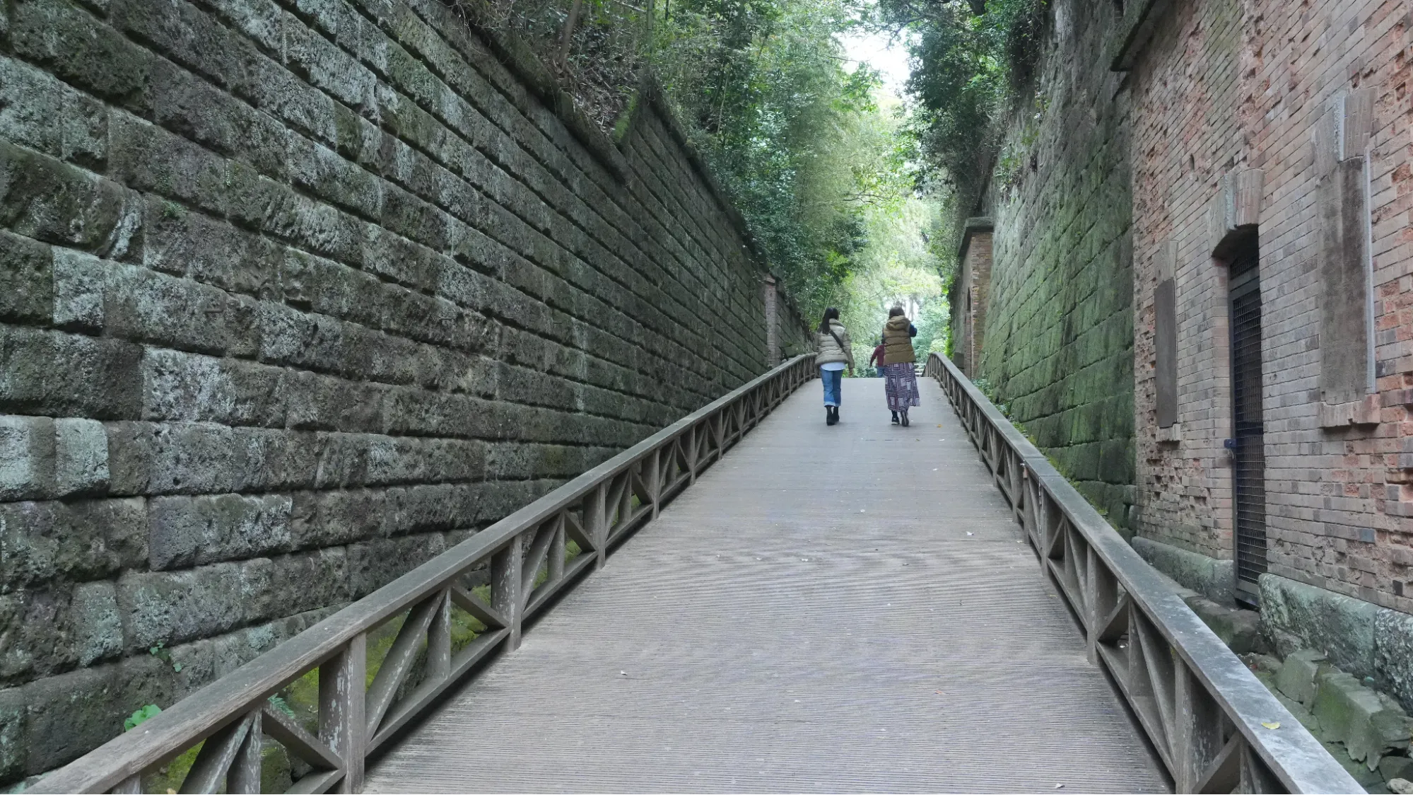兩個女生漫步在棧道上，棧道旁邊有一面磚牆