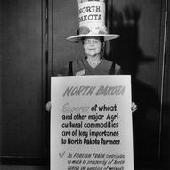 Woman wearing a tall “North Dakota” hat decorated with wheat holds a sign about wheat exports and foreign trade, standing indoors against a dark backdrop.