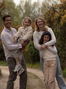 Allison family - A family modelling shot with two parents with natural expressions surrounded by their two children, their youngest daughter with short blonde hair and their eldest daughter with a wide bright smile, all together outside, represented by ROC Talent Agency UK.