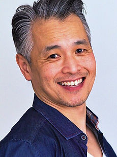 Nicky CH - headshot of a male actor with swept back hair, light stubble, and wearing a blue shirt, represented by ROC Talent Agency UK