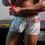 Thumbnail: Side view of male model in kitchen wearing colorful mini shorts with flirtatious cut outs on the sides.