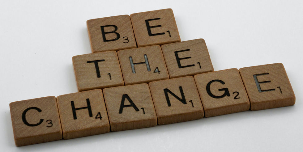 Scrabble tiles spell "BE THE CHANGE" on a white background. The wooden tiles have black letters and score numbers. Motivational mood.