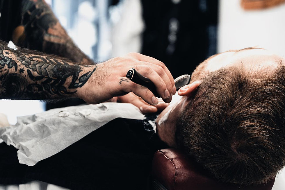 Tattooed barber shaves a man's neck with a straight razor. The client is reclined in a chair, covered with a white cloth. Calm ambiance.