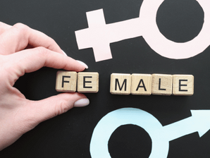 Main posant des blocs "FEMALE" sur fond noir avec symboles genre rose et bleu. Mains positionnant les blocs. Ambiance neutre et éducative.