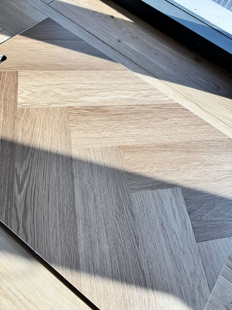 Sunlit close-up of light wood herringbone floorboards with strong diagonal shadows and a window edge at the top.