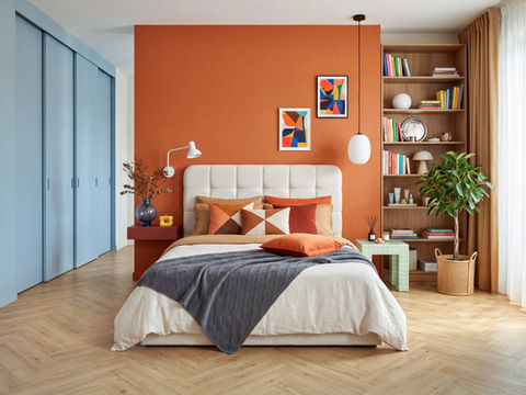 Modern bedroom with orange accent wall, cream bed, colorful pillows, built-in bookshelf, plant, and soft natural light