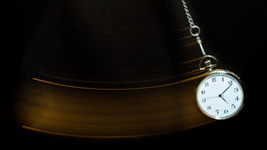 Pocket watch swinging on a chain against a dark background. Blurred motion trails suggest movement. White face with black numbers.
