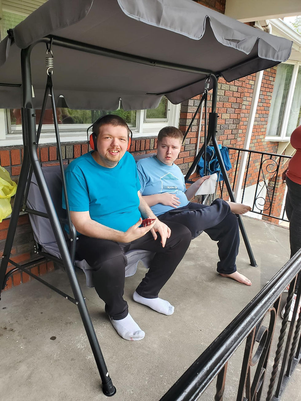 Two male residents at Penn Residential with intellectual disabilities sit on a porch swing together. Both wearing blue shirts and one of them wearing headphones, they look to the camera for a photo.
