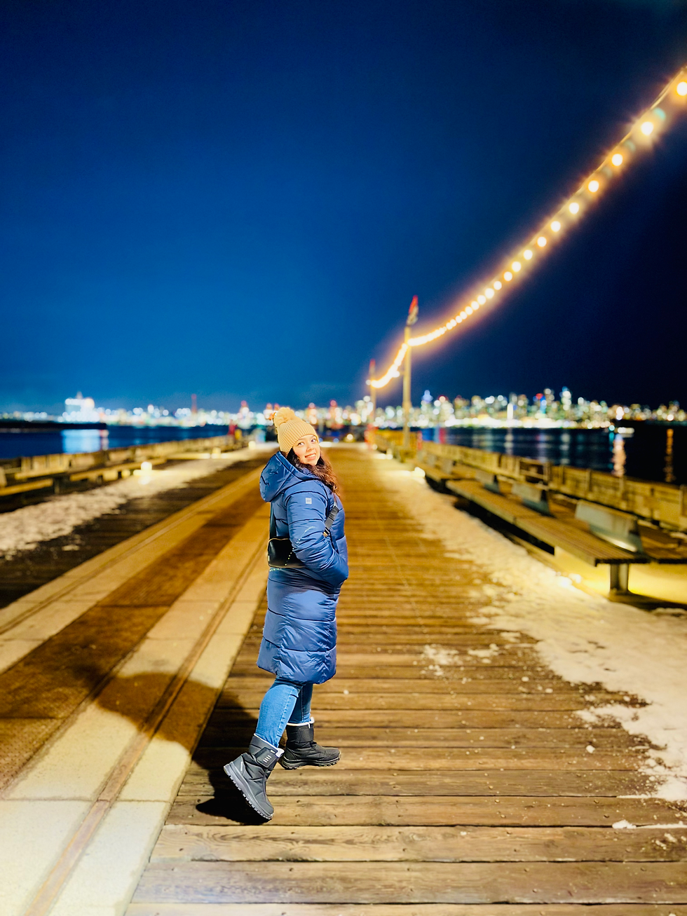 Person in a blue coat and beanie smiles on a snowy, wooden pier at night, with city lights and string lights in the background.