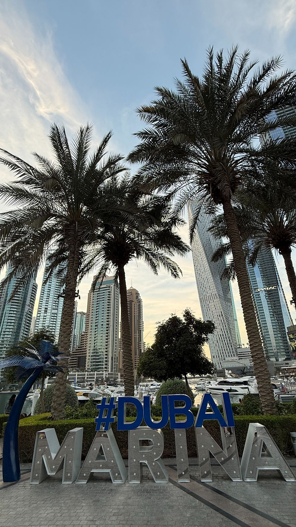 Palm trees and skyscrapers frame the "Dubai Marina" sign in blue and silver. Boats are docked, and a clear sky sets a tranquil mood.