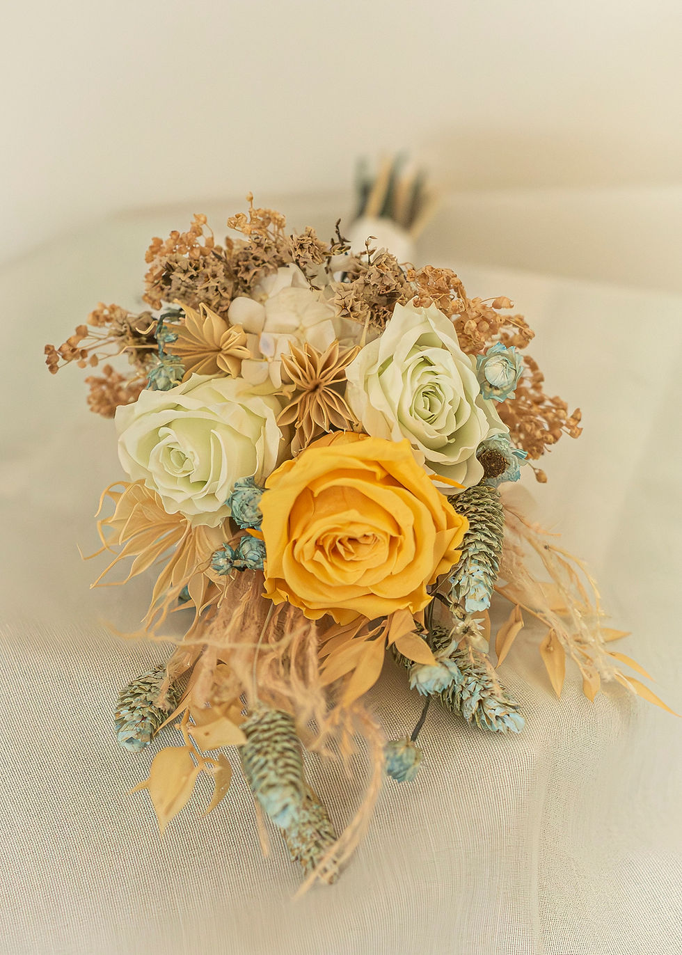 Preserved rose bridal bouquet with one golden yellow rose and two Blue Margarita roses, surrounded by tan dried flowers, feathery grasses and pale blue accents, lying on cream linen.