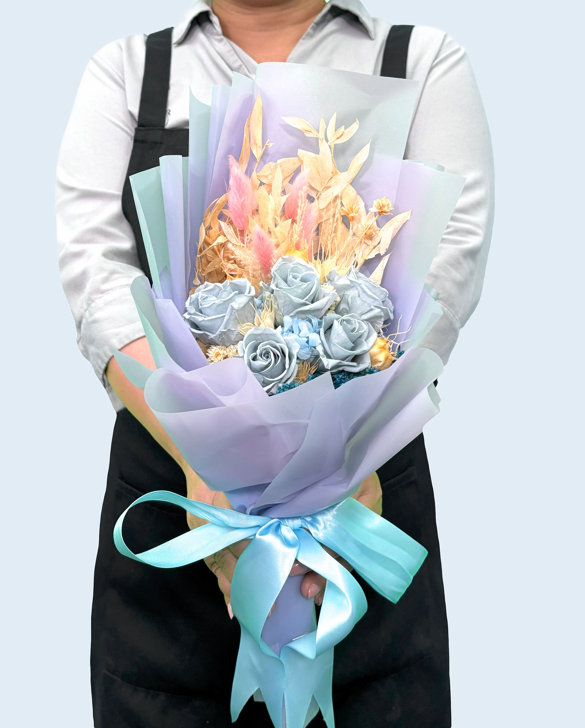 A person holding a beautiful SKfleur preserved bouquet featuring light blue roses mixed with pastel dried bunny tails, wrapped in soft purple paper with a bright blue ribbon.