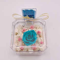 Close-up of a clear square acrylic bottle containing a preserved turquoise rose and small white and pink filler flowers, tied with a gold ribbon. This is a SKfleur 'Blooms in a Bottle' Christmas gift.