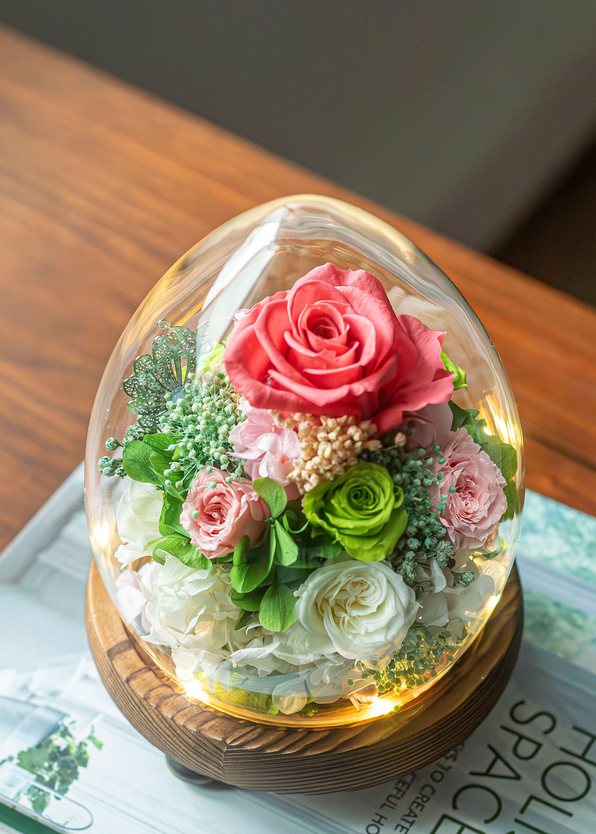 An elegant teardrop glass dome by SKfleur showcasing a large bright coral-red preserved rose, surrounded by smaller pink and lime-green roses with warm fairy lights on a wooden base.