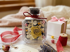 A square glass bottle with a preserved yellow rose and white flowers sits on a rustic wooden table next to a "For You" gift tag, dried lavender, and pink ribbon.