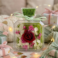 A square glass bottle with a red preserved rose and pink hydrangeas on a wooden table, styled with holiday gift boxes, pinecones, and warm fairy lights.