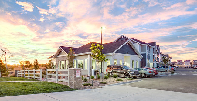 A modern apartment complex with a clubhouse, parking lot, and scenic sunset sky in the background.