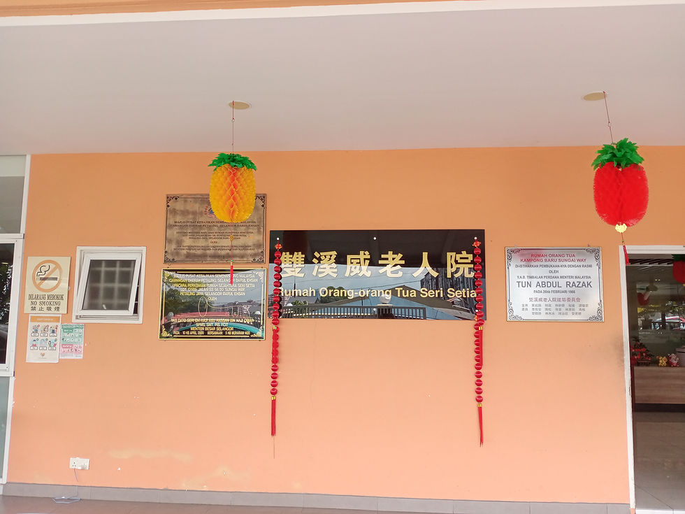 Orange building facade with bilingual signs for "Rumah Orang Tua Seri Setia." Hanging pineapple and strawberry decorations; "No Smoking" sign visible.