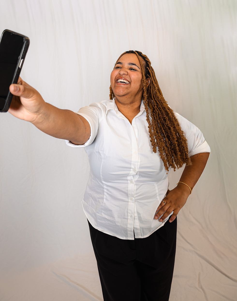 Young adult female (Jasmine Andrew) holds her iPhone up to take a picture of herself while smiling. She’s standing behind a white backdrop.