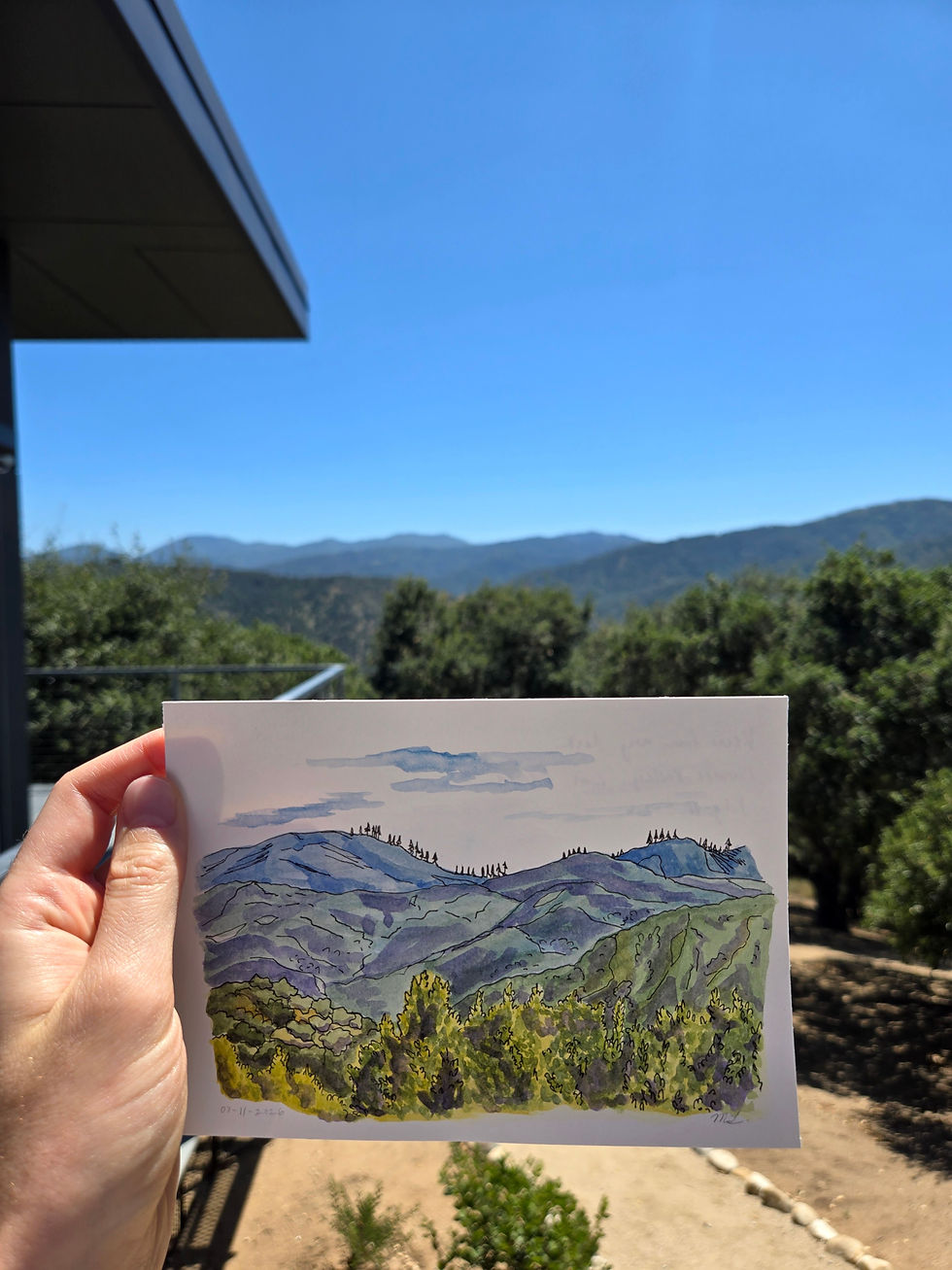 A hand holding up a 5 by 7 inch watercolor painting of blue hills beyond green treetops. The painting is being held up in front of the view that was painted.