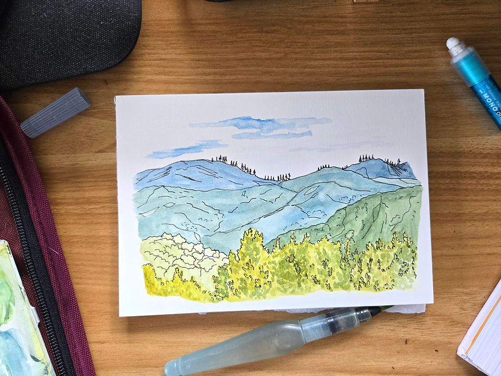A pad of watercolor paper with a pen and watercolor sketch of mountains with treetops in the foreground, sitting on a desktop with a water brush, eraser and the side of a watercolor palette.