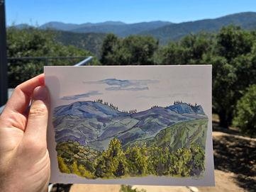 A hand holding up a 5 by 7 inch watercolor painting of blue hills beyond green treetops. The painting is being held up in front of the view that was painted.