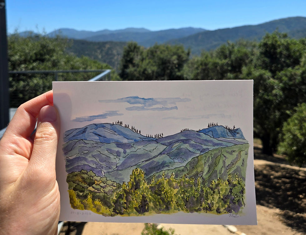 A hand holding up a 5 by 7 inch watercolor painting of blue hills beyond green treetops. The painting is being held up in front of the view that was painted.
