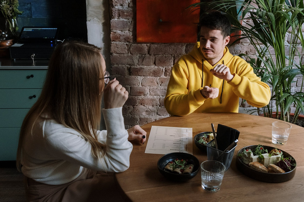 Two people sit at a table in a restaurant talking with sign language