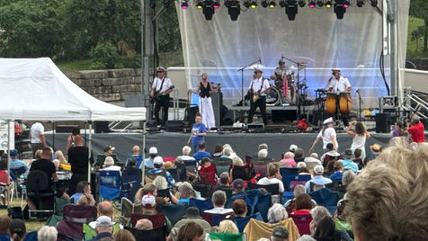 Band onstage at Olathe Live with large stage and white backdrop. Band dressed in white nautical uniforms and captain's hats. Large outdoor concert with audience and white tent
