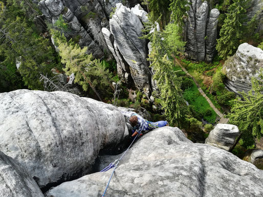 Lezec v široké spáře pod první kruh na věži v Adršpachu, v pozadí typické pískovcové věže a borovice.