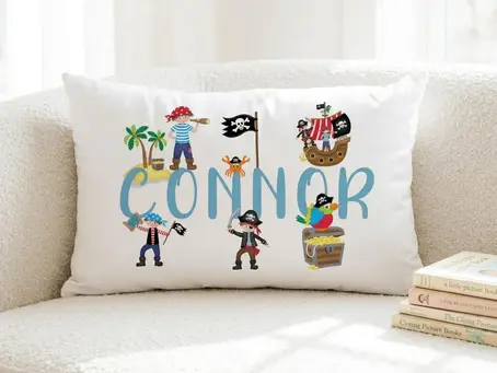 Personalised kids pirate pillowcase on a white textured couch next to a stack of children's books, featuring a blue custom name and pirate illustrations.