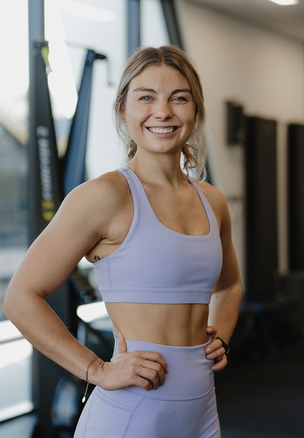 Female personal trainer in purple matching gym gear, smiling inside a gym with equipment around.