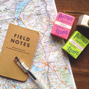 Field Notes notebook and pen on a colorful map. Nearby are pink, green, and white candy boxes. Wooden table background. Vintage travel vibe.