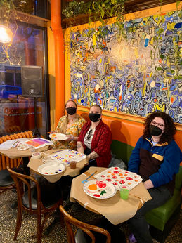 3 people sitting at a table at a cafe painting canvas tote bags