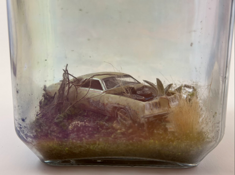 A 1/64 scale car set up in an abandoned scene. It is rusted and falling to pieces, with the hood slid underneath and a tire missing. Foliage is scattered around it and growing from the engine.