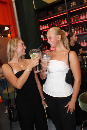 Zwei lächelnde Frauen stoßen mit Cocktails in einer modernen Bar an.
