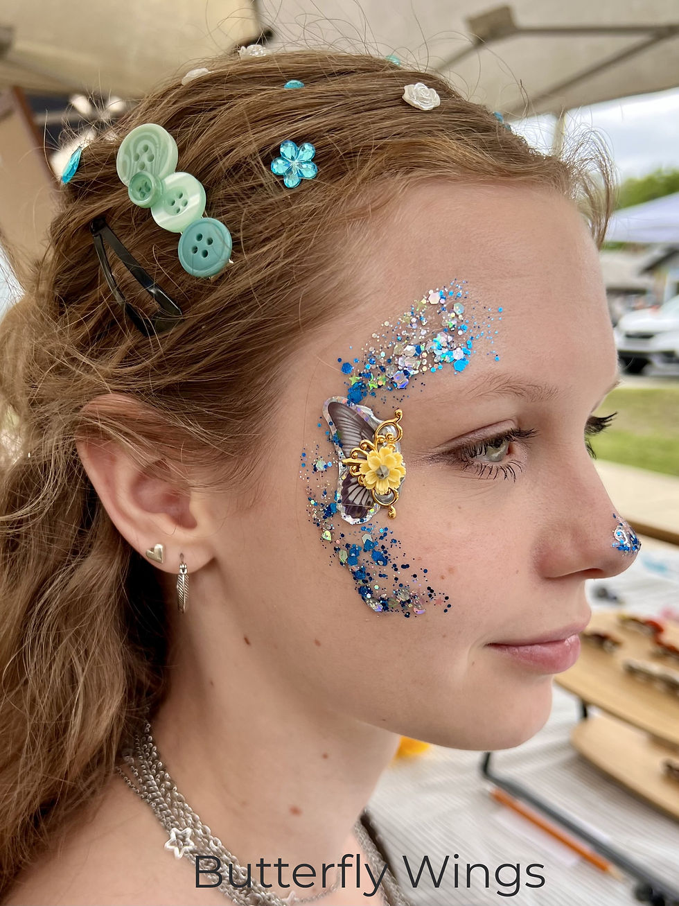 The image shows a side profile of a young child with light skin and blonde hair pulled back into a ponytail. The child is looking off to the side with a calm expression. A colorful butterfly-style face bling design decorates the area around one eye and cheek, featuring bright glitter colors such as pink, purple, and white with decorative swirls and dots. The background is blurred, keeping the focus on the child’s face and the face paint, suggesting a festive or recreational environment such as a fair or party.