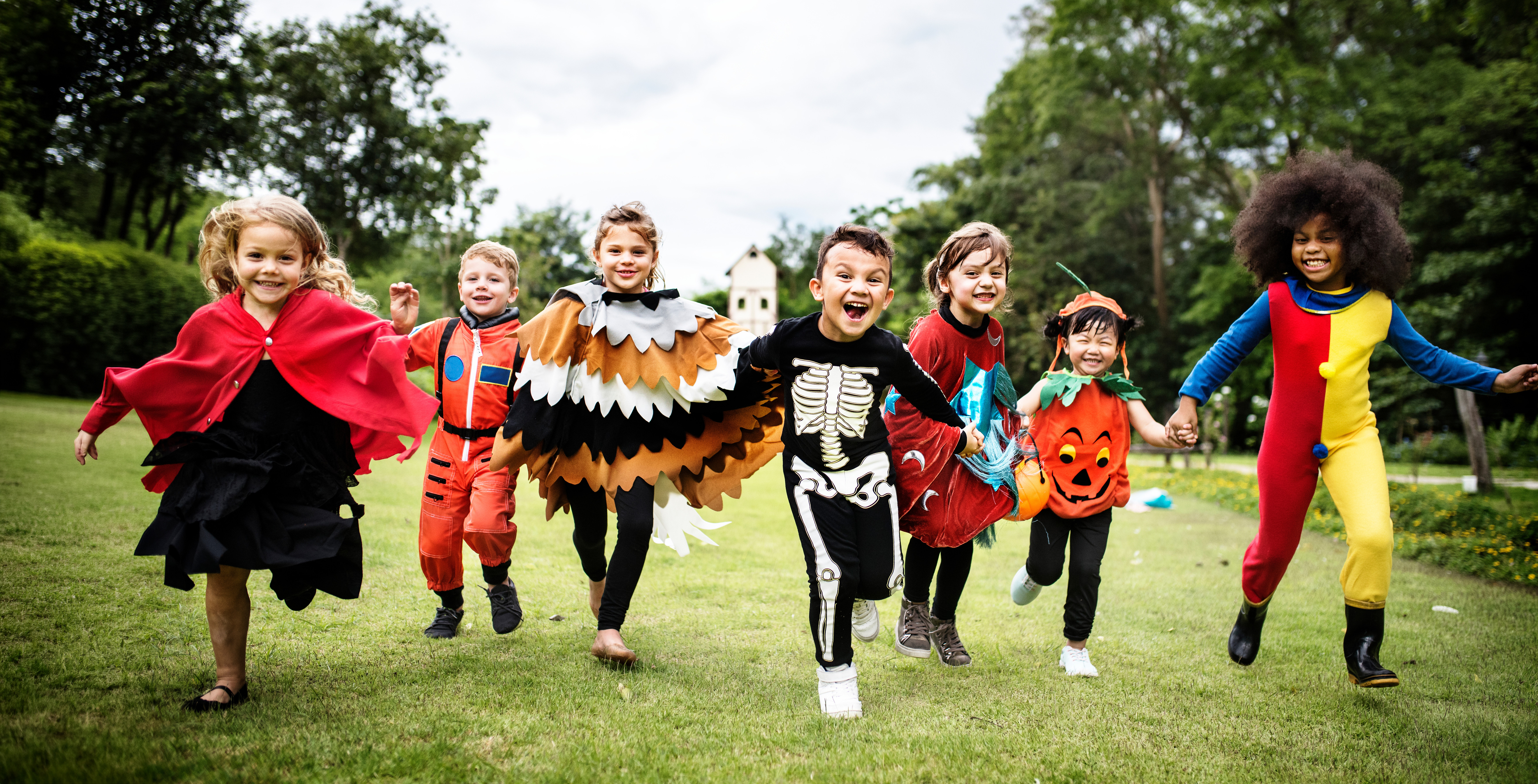Children in colorful Halloween costumes run across a grassy park, smiling and holding hands under a cloudy sky.