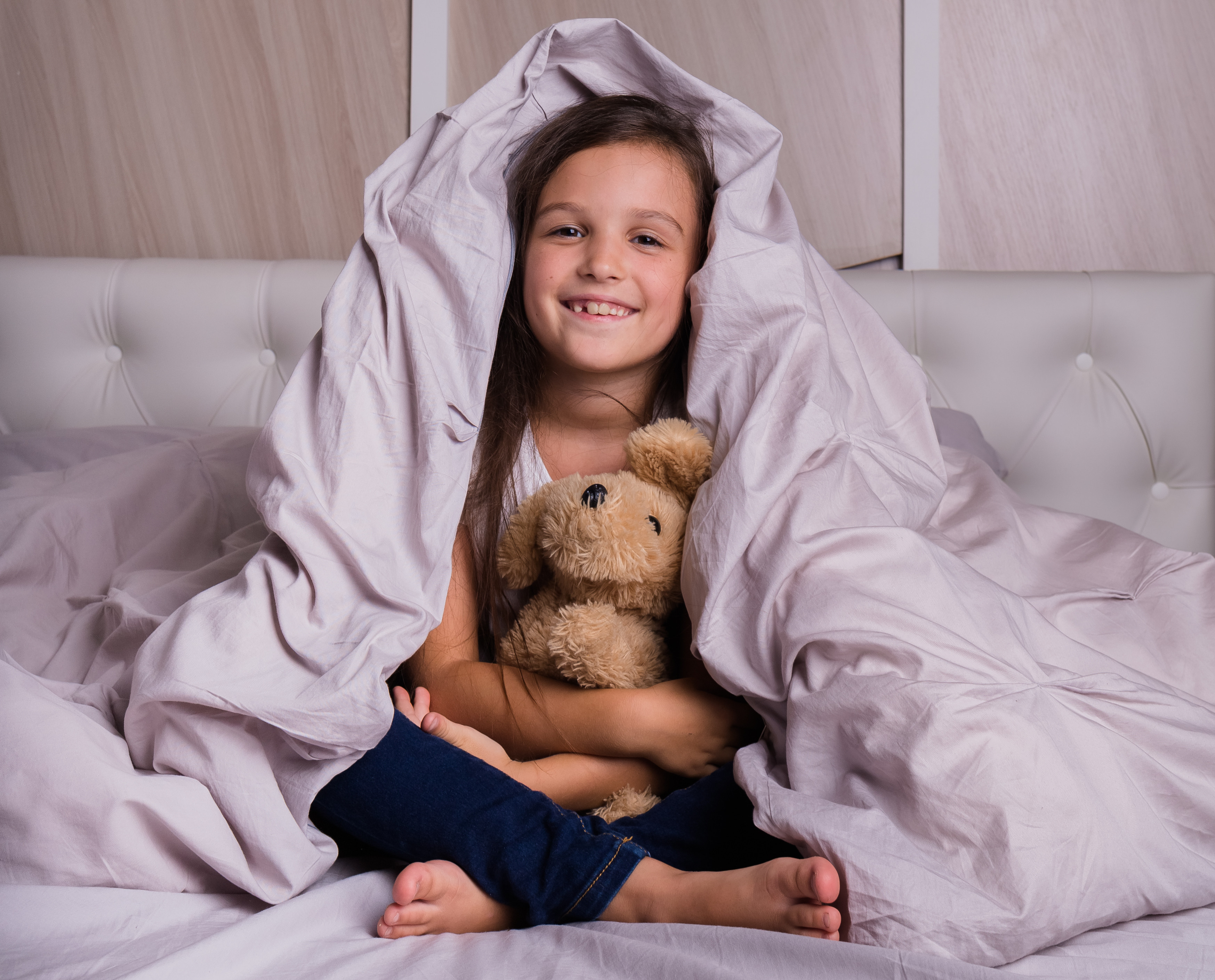 Smiling girl sits cross-legged in bed under a blanket, hugging a teddy bear in a cozy bedroom.