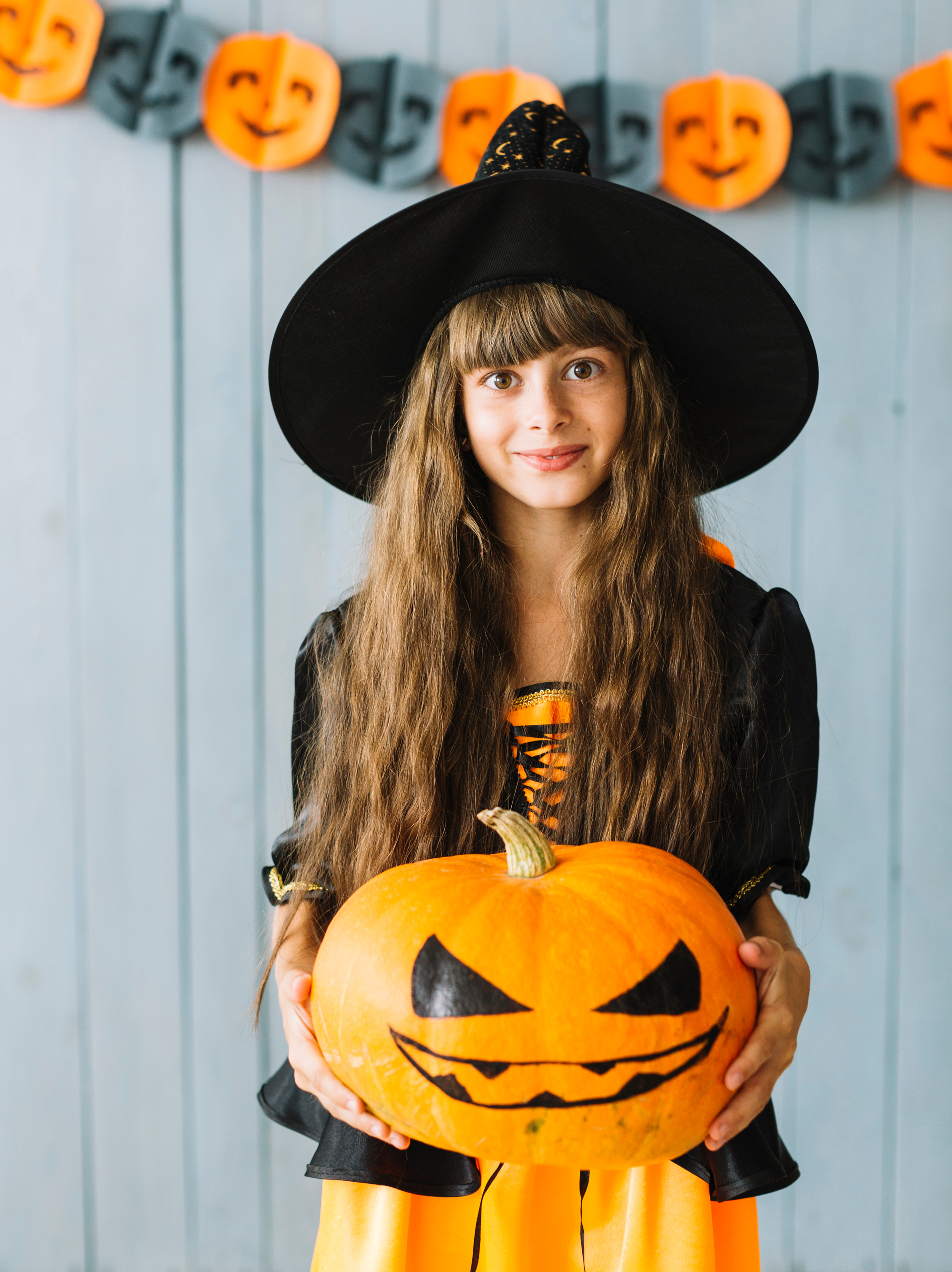 Girl in a witch costume holds a carved Halloween pumpkin, smiling in front of orange-and-gray smiling-face bunting.