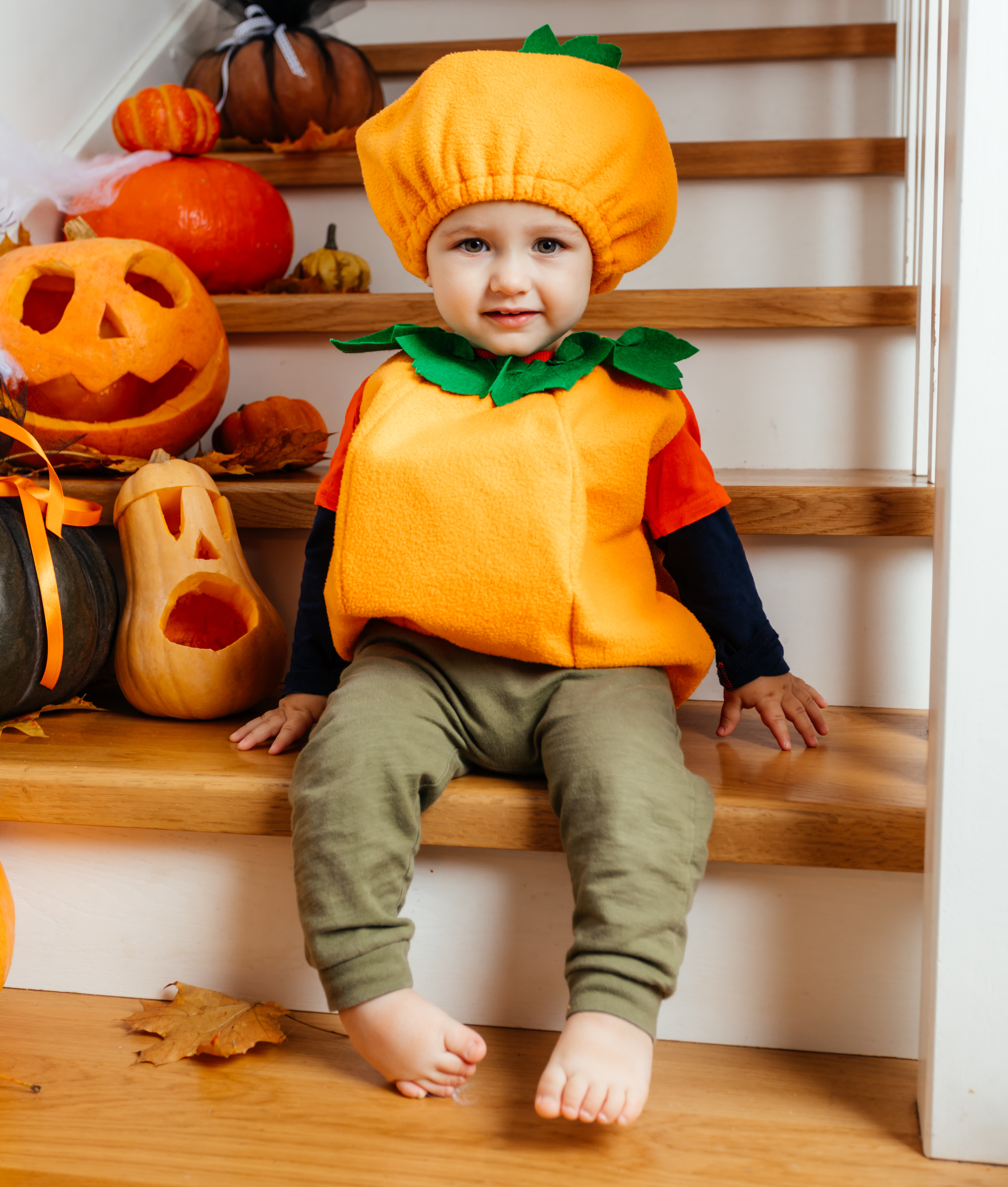 Toddler in a pumpkin costume sits on Halloween-decorated stairs beside carved pumpkins, looking calm and curious.