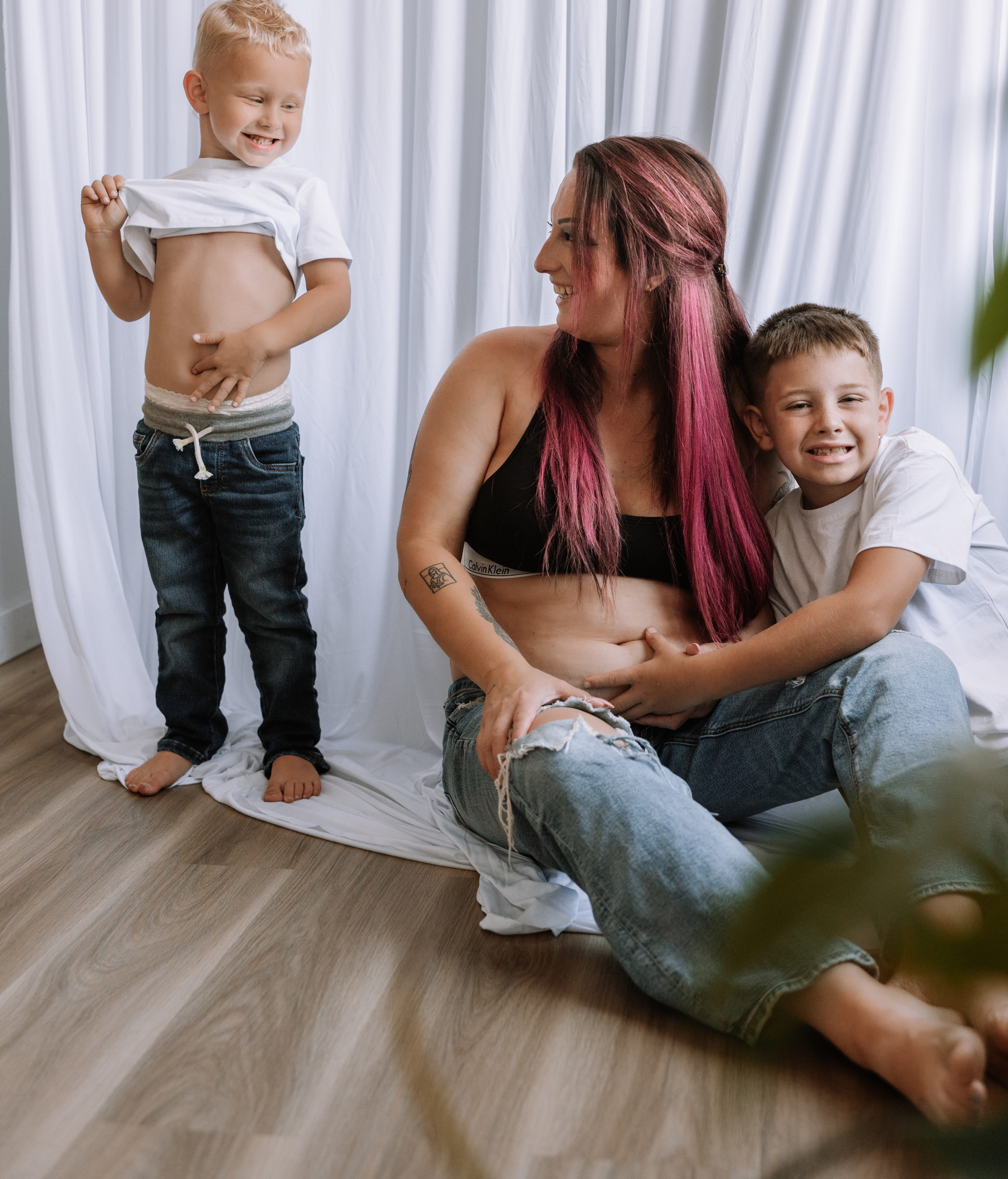 Smiling mother with pink hair and two young boys in a bright room with white curtains; one boy lifts his shirt, showing his belly.