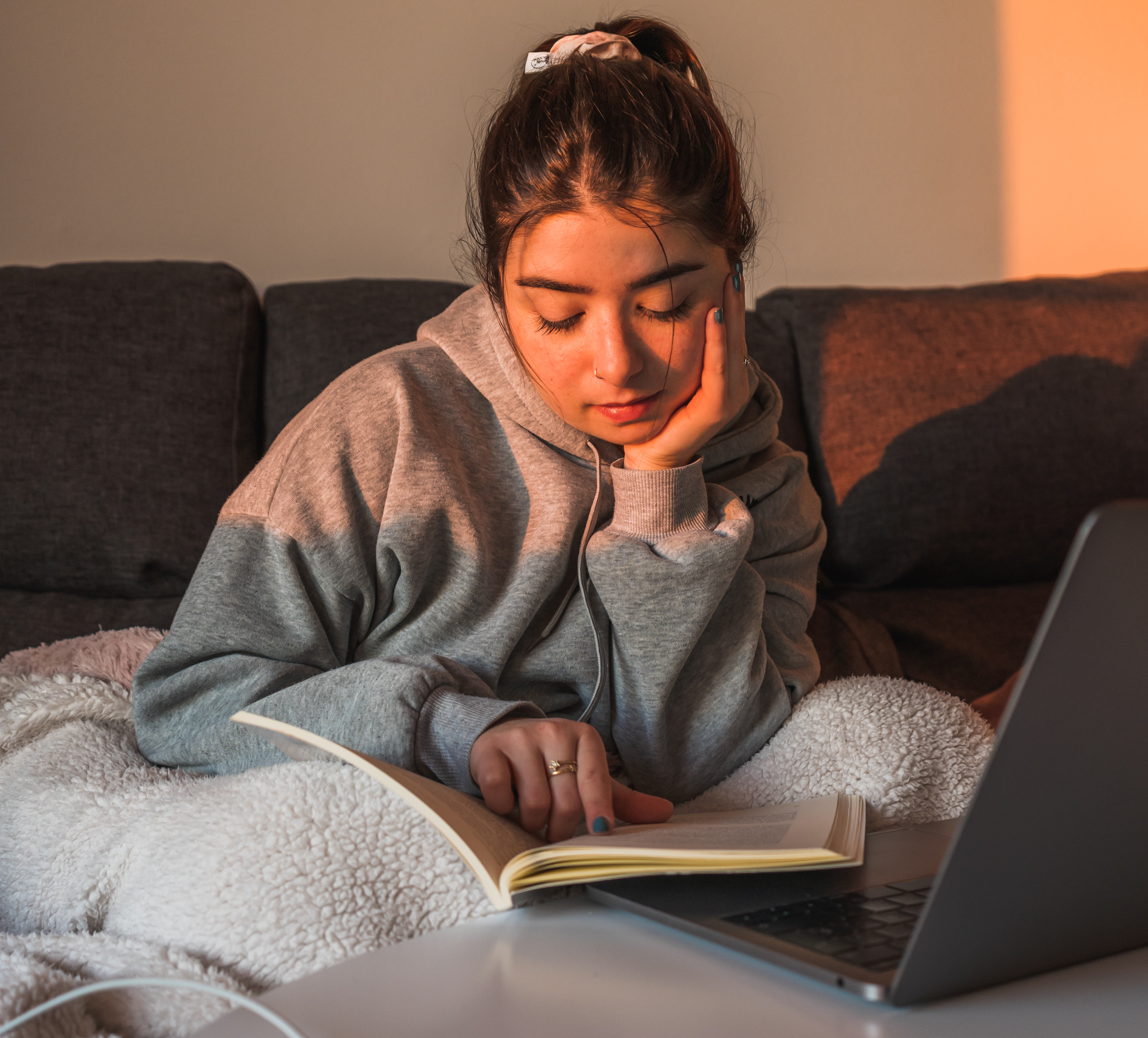 Young woman in a gray hoodie reads a book on a couch beside a laptop, bathed in warm sunlight.